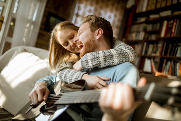 Hombre tocando la guitarra acústica en el sofá para su joven bella mujer