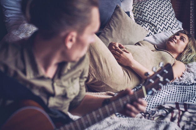 Hombre tocando una guitarra y su esposa embarazada están acostados en una cama en una habitación tipo loft