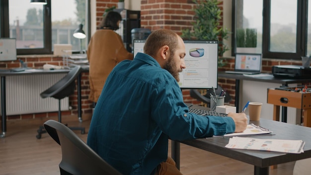 Hombre tomando notas sobre papeles gráficos y mirando la pantalla del ordenador. Empleado de negocios que trabaja con datos e información en el monitor, escribiendo en archivos para planificar el proyecto y la presentación.