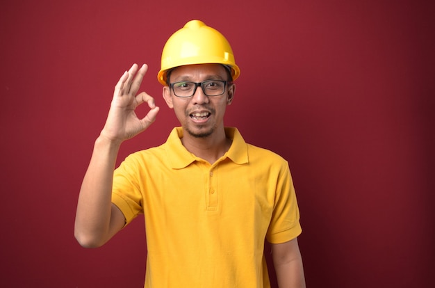 Hombre trabajador asiático guapo con casco sonriendo y muestra gesto bien contra el fondo rojo.