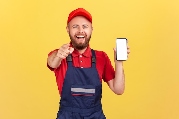 Hombre trabajador con camiseta roja de overol apuntándote con el dedo y mostrando un teléfono inteligente con pantalla en blanco