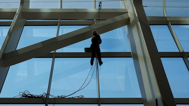 Hombre trabajador sin rostro está lavando ventanas con un raspador en un edificio moderno de gran altura colgando la cuerda.