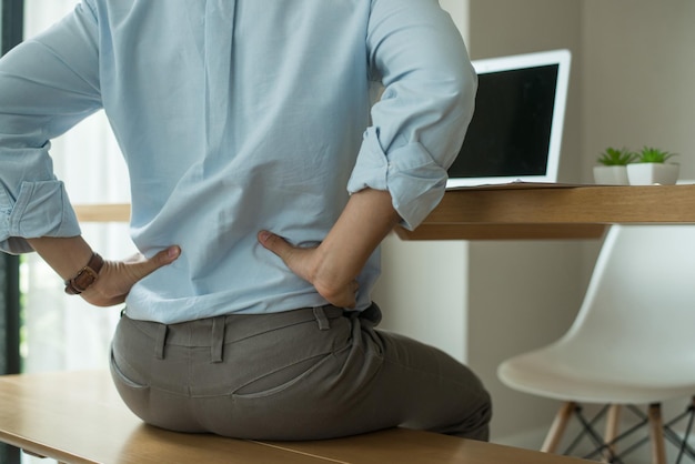 Hombre trabajador sentado en la silla del banco de madera con una computadora portátil en el escritorio en la sala de la oficina Usó ambas manos para presionar la parte inferior de la espalda Sufre dolor de espalda por estar sentado durante mucho tiempo