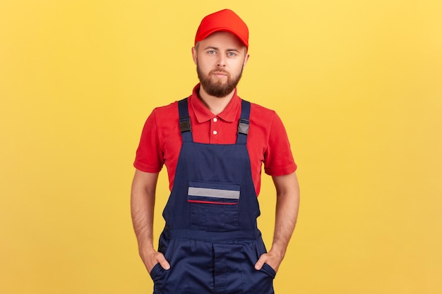 Hombre trabajador con uniforme azul y gorra roja de pie con las manos en el bolsillo