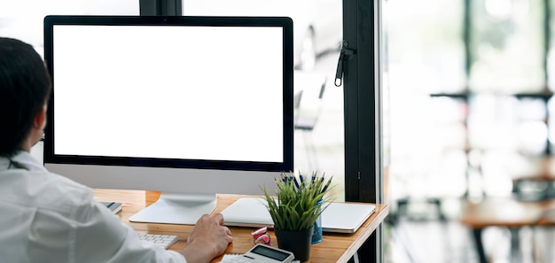 Hombre trabajando en una computadora de escritorio en casa, maquetando un monitor de pantalla en blanco para mostrar productos o diseño gráfico. Fondo panorámico de la oficina.