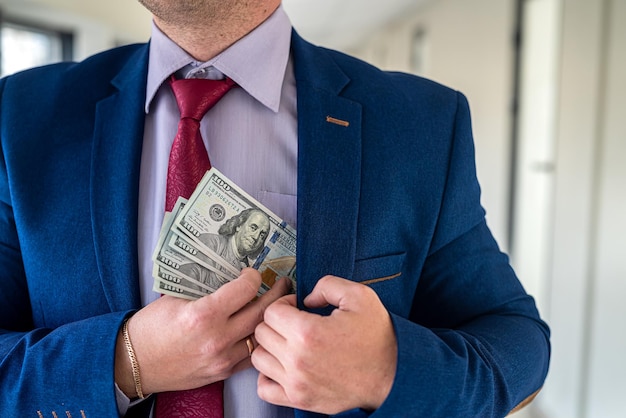 Hombre de traje azul y corbata roja poniendo billetes de dólar de soborno en su concepto de corrupción de bolsillo