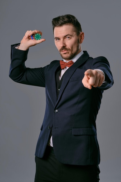 Hombre con traje clásico negro y corbatín rojo que muestra algunas fichas de colores posando contra el fondo gris del estudio...