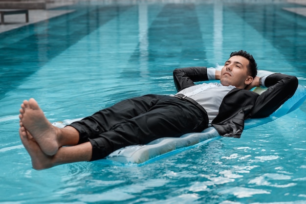 Un hombre con un traje está descansando en la piscina sobre un colchón azul inflable