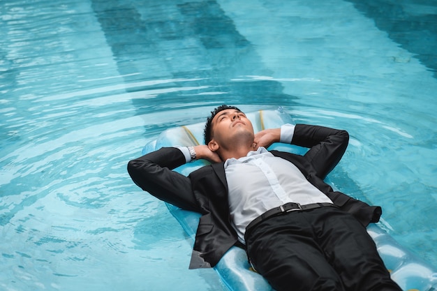 Un hombre con un traje está descansando en la piscina sobre un colchón azul inflable