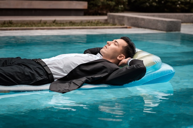 Un hombre con un traje está descansando en la piscina sobre un colchón azul inflable