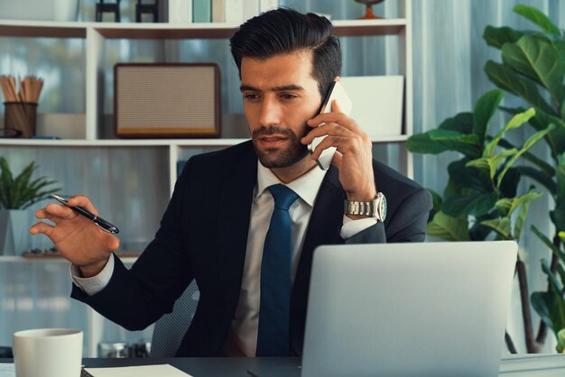 Un hombre con traje está hablando por teléfono y sostiene un bolígrafo y una computadora portátil.