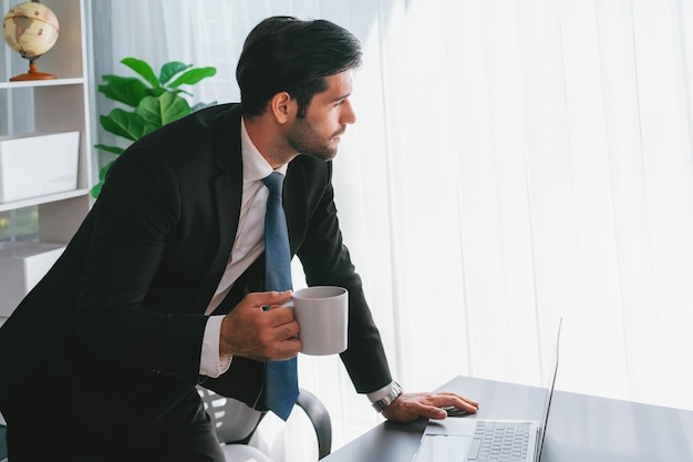 Un hombre con traje está mirando una computadora portátil y sosteniendo una taza.