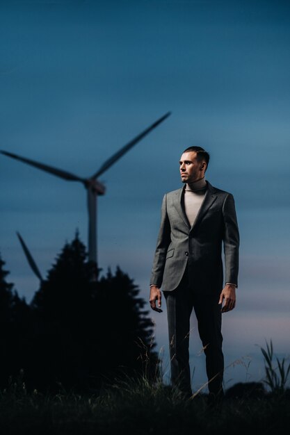 Un hombre con un traje gris se encuentra junto a un molino de viento después del atardecer. Hombre de negocios cerca de los molinos de viento en la noche. Concepto moderno del futuro.