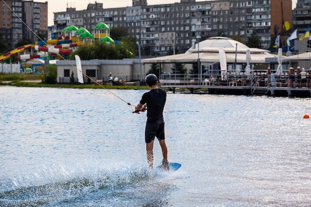 Hombre en traje húmedo y casco protector sosteniendo cuerda de remolque y balanceándose en wakeboard