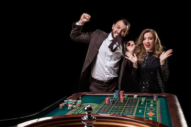 Un hombre con un traje marrón con una mujer hermosa con un vestido negro está jugando en una mesa de ruleta en un casino. Emoción de los jugadores de juego