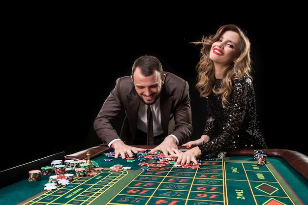 Un hombre con un traje marrón con una mujer hermosa con un vestido negro está jugando en una mesa de ruleta en un casino. Emoción de los jugadores de juego