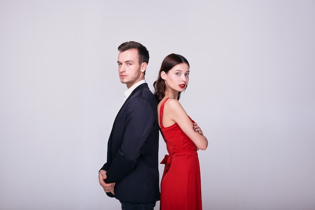 Hombre en traje y mujer en vestido rojo espalda con espalda sobre fondo blanco.