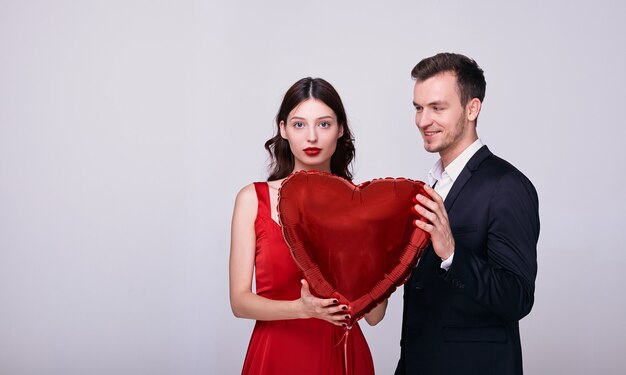 El hombre en traje y la mujer en vestido rojo sostienen un globo en forma de corazón rojo sobre fondo blanco.