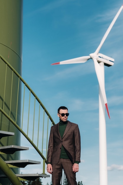 Un hombre en un traje de negocios con una camisa de golf verde se encuentra junto a un molino de viento contra el fondo del campo y el cielo azul. Hombre de negocios cerca de los molinos de viento. Concepto moderno del futuro.