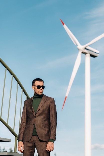 Un hombre en un traje de negocios con una camisa de golf verde se encuentra junto a un molino de viento contra el fondo del campo y el cielo azul. Hombre de negocios cerca de los molinos de viento. Concepto moderno del futuro.