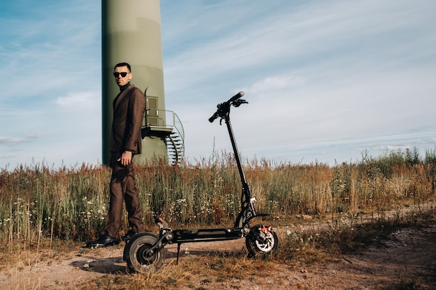 Un hombre con un traje de negocios y una camisa de golf verde se encuentra junto a un scooter eléctrico contra un campo y un cielo azul. Hombre de negocios cerca del scooter. Concepto moderno del futuro.