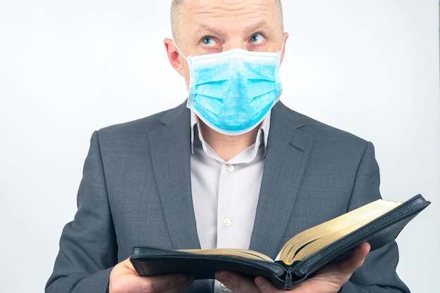 El hombre en un traje de negocios con una máscara médica en su rostro está estudiando la Biblia.