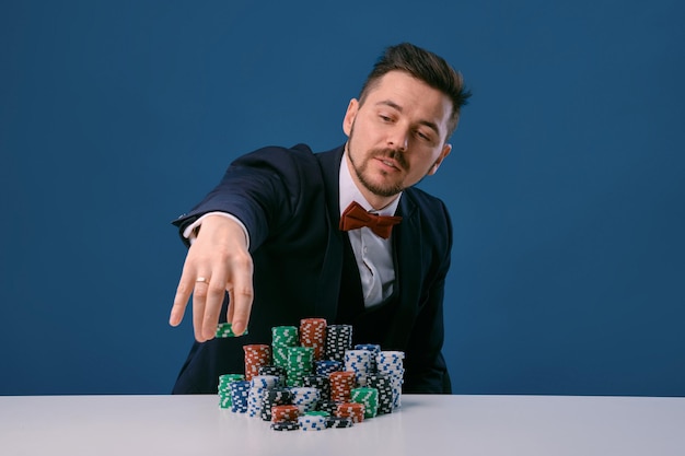 El hombre de traje negro está sentado en una mesa blanca con montones de fichas de colores posando en un estudio azul...