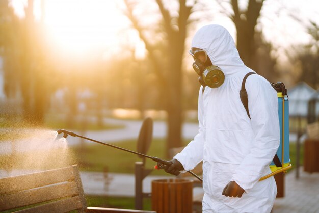 Hombre en traje protector desinfectando banco en el parque público en la ciudad de cuarentena. Prevención de infecciones y control de epidemias. COVID-19.