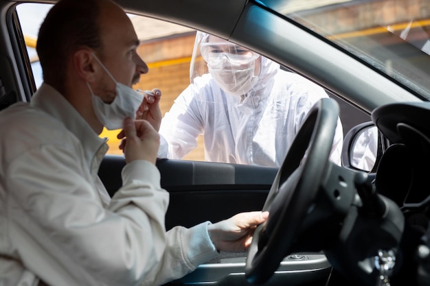 Un hombre con traje protector, guantes, mascarilla quirúrgica y mascarilla está probando el coronavirus co id-19 en otro hombre sentado en un automóvil.