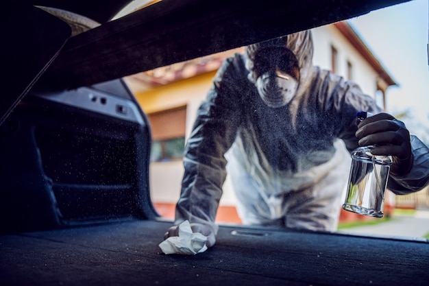 Hombre con traje protector con máscara desinfectando el interior del coche, limpiando las superficies que se tocan con frecuencia, previene la infección por coronavirus, la contaminación de gérmenes o bacterias. Infección