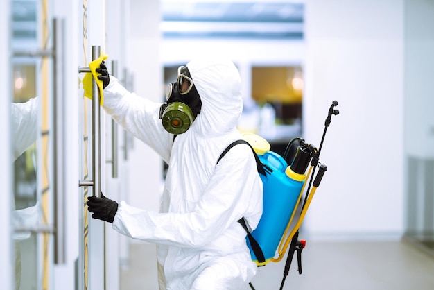 El hombre con traje protector de materiales peligrosos lava los muebles de oficina para prevenir la propagación del coronavirus.