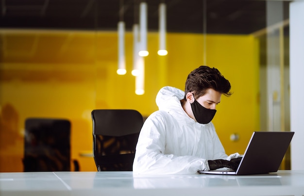 El hombre con traje protector de materiales peligrosos y máscara trabaja en una computadora en una oficina vacía.