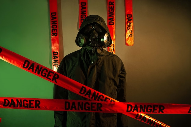 Un hombre con un traje protector oscuro con una máscara de gas en la cara y una capucha en la cabeza posando de pie cerca de una pared verde con una cruz de cintas de peligro. Concepto de peligro