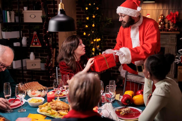 Hombre en traje de santa claus saludando a mujer, dando regalo de navidad, esposa recibiendo caja de regalo de marido. Celebración de la noche de Navidad, gran familia diversa comiendo comida tradicional en la mesa festiva