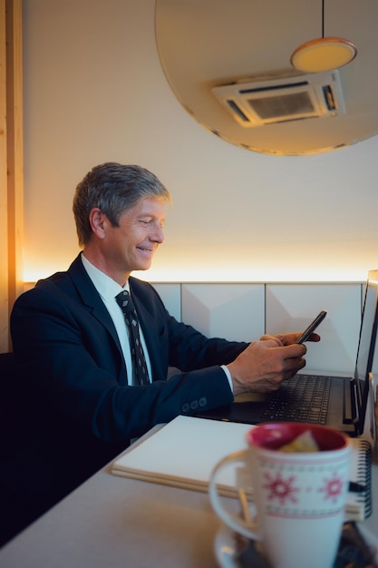 Hombre de traje sentado en la mesa con una computadora portátil mirando su teléfono móvil