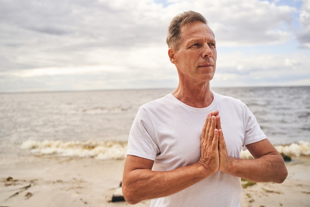 Hombre tranquilo está de pie en la playa y practica yoga mientras se sostiene de la mano en posición namaste