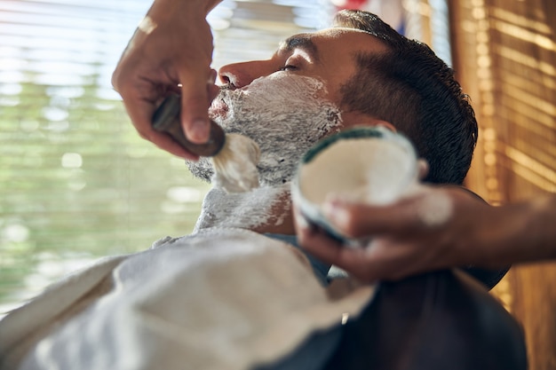 Hombre tranquilo con los ojos cerrados cubiertos de crema de afeitar durante una cita en una barbería