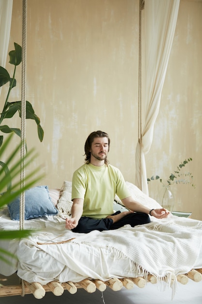 Hombre tranquilo sentado en la cama y haciendo meditación matutina, profesor de yoga en casa de diseño moderno con plantas verdes practicando yoga