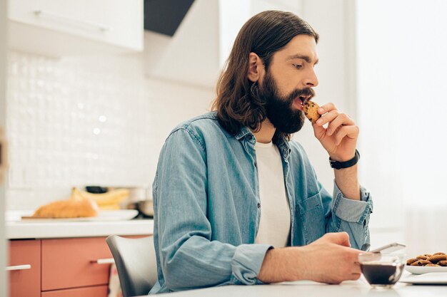 Hombre tranquilo con un teléfono inteligente sentado en la mesa y mordiendo una galleta. Taza de café a su lado