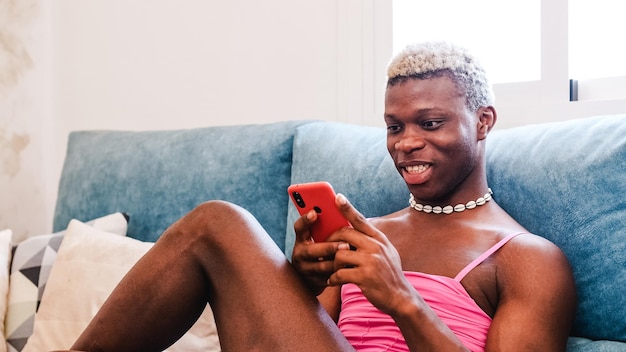 Hombre transexual negro africano con ropa femenina elegante enviando mensajes de texto en el teléfono celular en el sofá