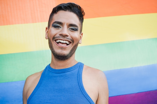 Foto hombre transgénero joven con maquillaje sonriendo a la cámara con la bandera del arco iris lgbt en el fondo - centrarse en la cara