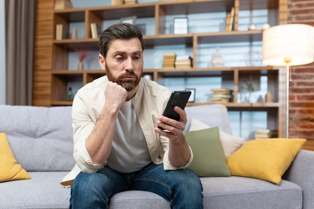 Hombre triste y deprimido solo en casa sentado en un sofá independiente con ropa informal sosteniendo un teléfono inteligente