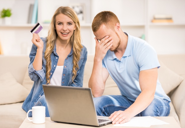 Hombre triste que trabaja con el ordenador portátil mientras que esposa que sostiene la tarjeta de crédito.