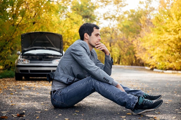 Un hombre triste se sienta en la carretera frente a un auto roto con el capó abierto