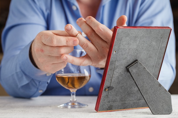 Foto hombre triste sostiene un anillo de bodas cerca del vaso de bebida alcohólica mientras está sentado en la cocina, primer plano