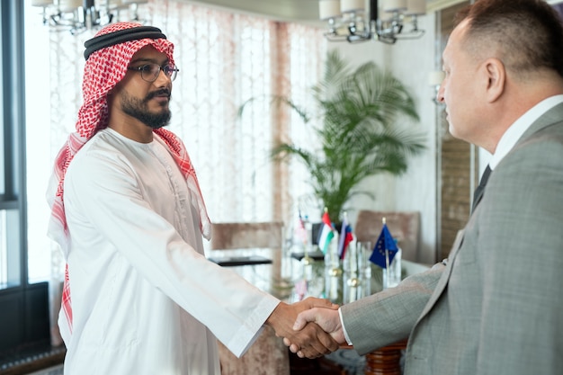 Foto un hombre con una túnica blanca un apretón de manos con un hombre con traje gris después de negociar
