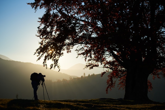 Hombre de turismo excursionista con cámara en el valle cubierto de hierba en el fondo del paisaje de montaña bajo un gran árbol.