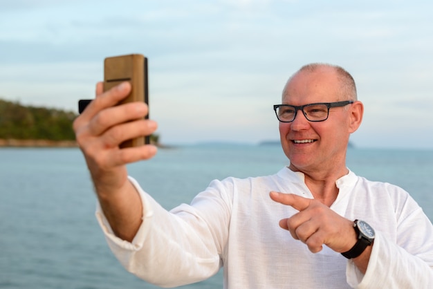 Hombre turista guapo maduro feliz tomando selfie contra la vista de la playa al aire libre