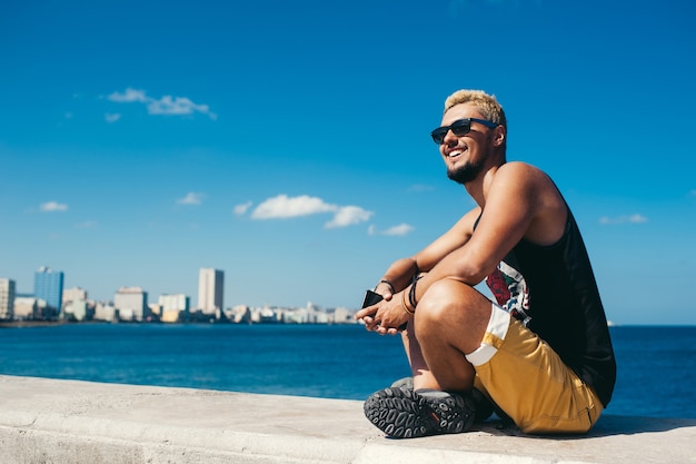 Hombre turista sentado en piedra mientras sonríe y posando contra el mar azul