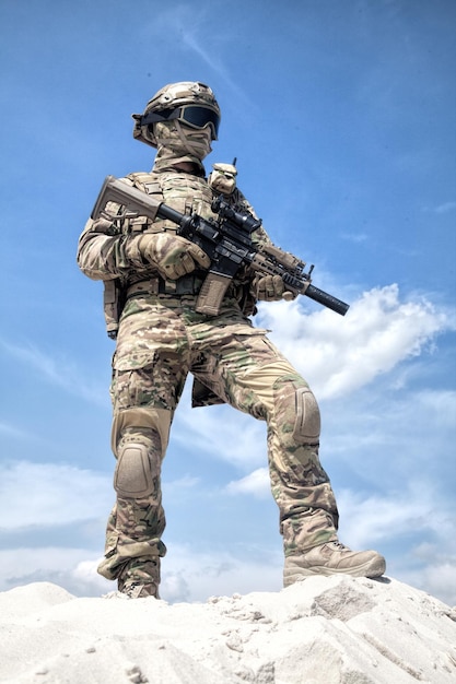 Hombre con uniforme de camuflaje militar y máscara, munición táctica equipada, de pie sobre una duna de arena con réplica de rifle de servicio en las manos, cielo nublado en el fondo. Jugador de airsoft participando en juegos de guerra